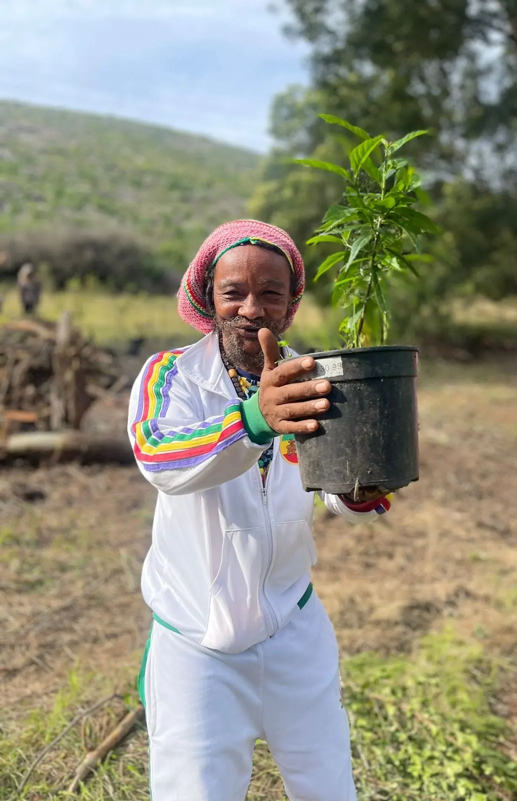 A man in a knitted cap smiles while holding a potted sapling